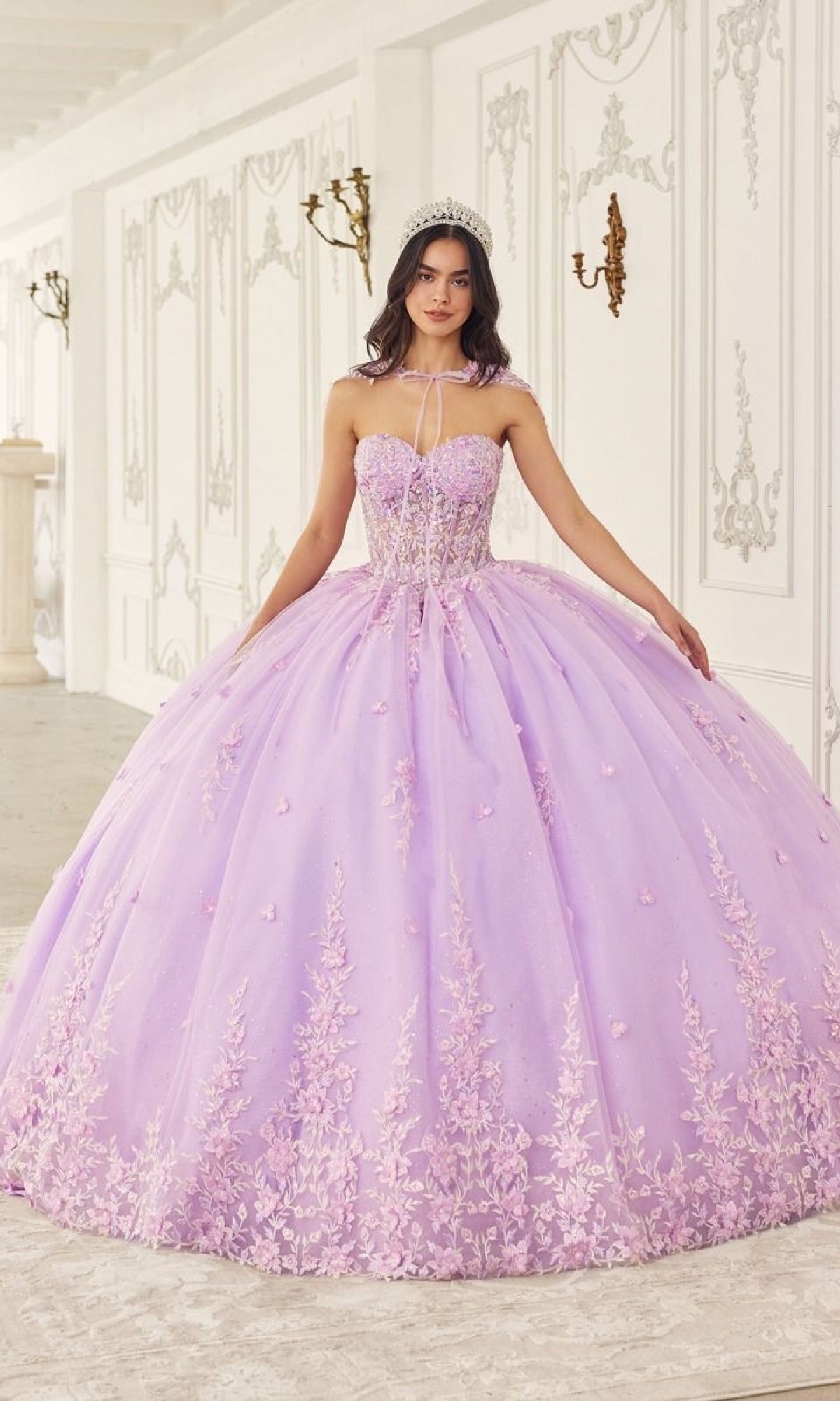 A young woman stands indoors wearing the Quinceanera Dress 15723 by Ladivine, a strapless lavender gown with a corset bodice, intricate embroidery, and floral lace applique, completed with a tiara in an elegant room.
